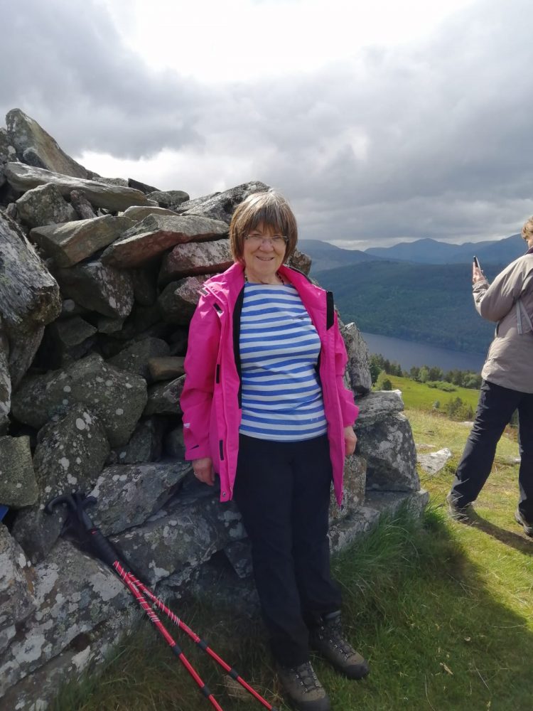 Woman wearing a blue and white striped top and pink jacket stands next to a stone cairn.