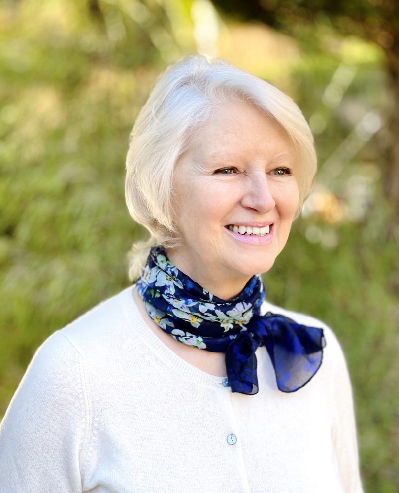 Woman with white hair wearing a white top and blue floral neckscarf.