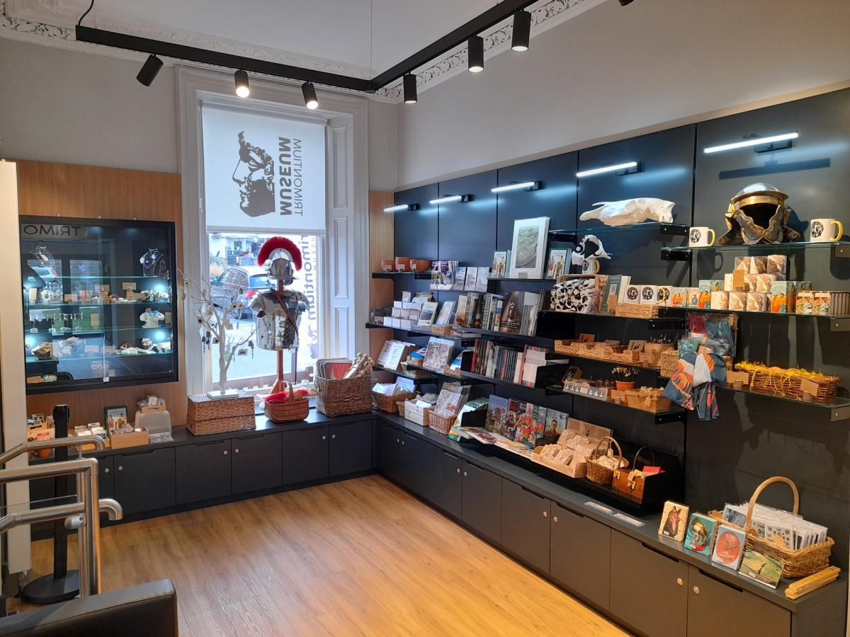 An L-shaped museum shop, including Trimontium mugs, cards, books, and other stock stored in wicker baskets and on black shelves.