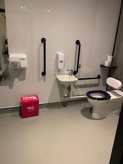View of an accessible toilet. A white toilet with black seat on the right hand side is next to a sink and black handrails. A hand dryer and Biffy bin are on the left.