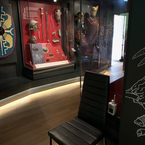 A black seat in the museum gallery with Roman military artefacts in a display case behind.