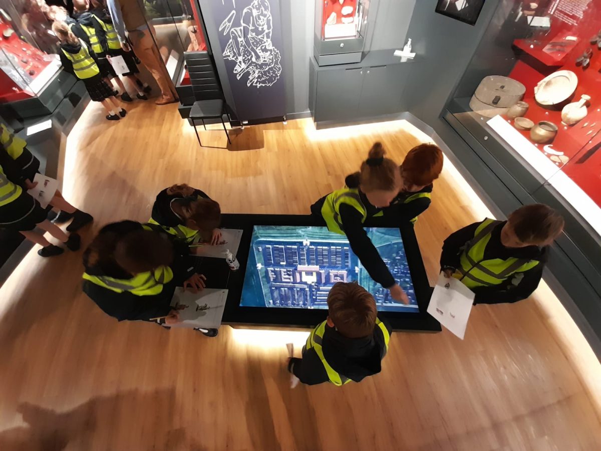 Children wearing hi-vis vests interact with a flat computer screen in the museum gallery.