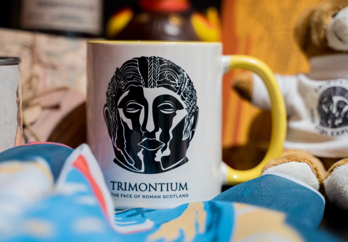 A white mug with yellow handle with the Trimontium logo in the centre.
