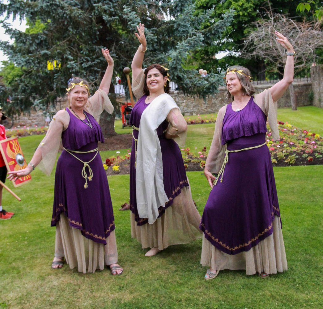 Three woman in purple ancient Roman-inspired dresses strike a pose.