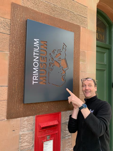 Man standing and pointing at the Trimontium Museum sign outside the museum.