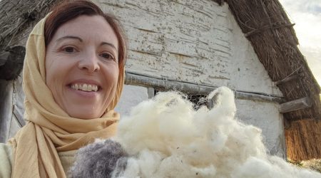 A woman in Carolingian clothes appears smiling in front of a house and holding wool.