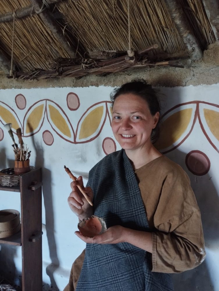 a woman in Iron Age costume inside an Iron Age dwelling holding paints and pigments